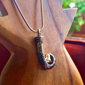 May include: A silver-toned pendant necklace featuring a stylized hockey stick design. The pendant has intricate etched details and a curved shape with a small, open circle at the end. The necklace is displayed on a wooden stand.