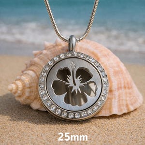 May include: A silver-toned pendant necklace with a round, crystal-studded frame. The pendant features a cut-out hibiscus flower design. The necklace is displayed on a sandy beach with a seashell in the background. The pendant is 25mm in diameter.