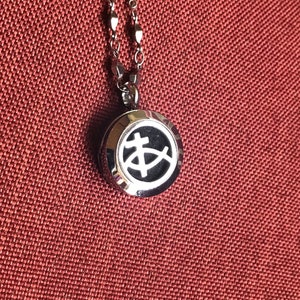 May include: A silver-colored necklace with a round pendant featuring a Christian fish symbol. The pendant has a black background and a silver outline. The necklace is displayed on a reddish-brown textured surface.