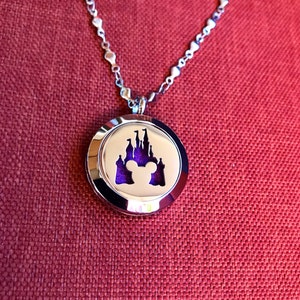 May include: A silver-toned necklace with a round pendant featuring a purple background and a white silhouette of a castle and Mickey Mouse ears. The necklace has a delicate chain and is displayed on a red textured surface.