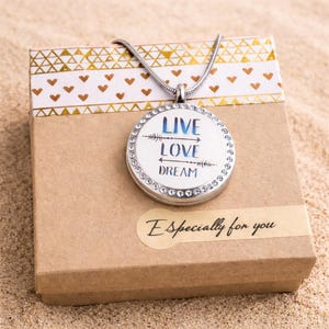 May include: A silver pendant necklace with a round charm that reads "LIVE LOVE DREAM" in blue and white. The charm is surrounded by small, clear crystals. The necklace rests on a brown gift box with a gold and white patterned top and a sticker that says "Especially for you."