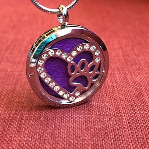 May include: A silver-colored pendant necklace with a round, openwork design. The pendant features a heart shape with a paw print cutout, embellished with clear crystals. The center has a purple insert. The necklace hangs on a silver chain.