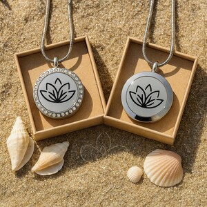 May include: Two silver necklaces with round pendants featuring a black lotus flower design, presented in small brown boxes. One pendant is adorned with clear crystals. The scene includes seashells on a sandy surface.