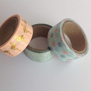 May include: Three rolls of washi tape. One roll has a pink background with gold flamingo silhouettes. Another roll has a light blue background with pink and blue floral patterns. The third roll has a silver glitter background.