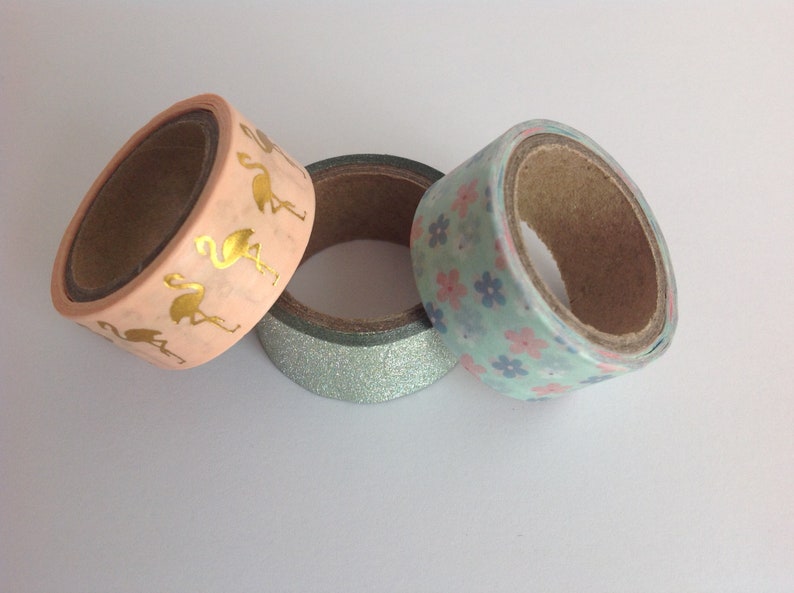 May include: Three rolls of washi tape. One roll has a pink background with gold flamingo silhouettes. Another roll has a light blue background with pink and blue floral patterns. The third roll has a silver glitter background.
