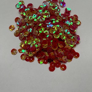 Könnte beinhalten: Ein Haufen von schimmernden roten Pailletten mit einem goldenen Schimmer. Die Pailletten sind klein und rund und auf einer weißen Oberfläche verstreut.