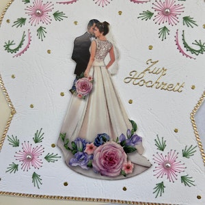 Puede incluir: Una tarjeta blanca con un diseño en forma de corazón. La tarjeta presenta una pareja con atuendo de boda, rodeada de flores bordadas en rosa y verde. El texto "Zur Hochzeit" está escrito en letras doradas.