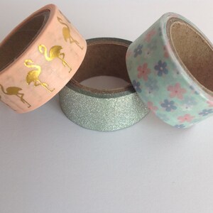 May include: Three rolls of washi tape. One roll has a pink background with gold flamingo silhouettes. Another roll has a light blue background with pink and blue flower silhouettes. The third roll has a light blue glitter background.