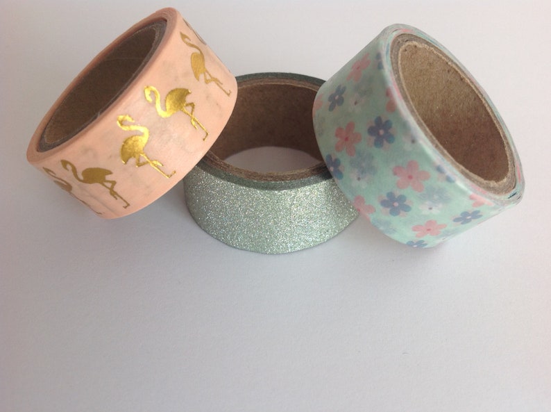 May include: Three rolls of washi tape. One roll has a pink background with gold flamingo silhouettes. Another roll has a light blue background with pink and blue flower silhouettes. The third roll has a light blue glitter background.