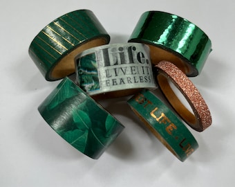Op de afbeelding: Assortiment van groene en gouden washi tape rollen. De rollen hebben verschillende patronen, waaronder strepen, bladeren en metallic afwerkingen. Eén rol heeft de tekst "Life. Live it Fearless" erop gedrukt.
