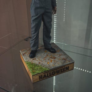 May include: A figure of a person in a gray jumpsuit and black shoes stands on a square base. The base has a concrete-like surface with patches of green grass and fallen leaves. The base is bordered by a wooden frame with the text "JOHN CARPENTER'S HALLOWEEN".