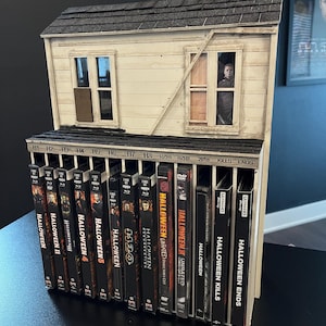 Puede incluir: Un estante de almacenamiento multimedia de madera con forma de casa, que presenta un techo de tejas y dos ventanas. El estante contiene una colección de cajas de películas de Halloween etiquetadas de H1 a H8, RZH1, RZH2, 2018, Kills y Ends.