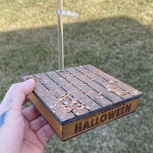 May include: A wooden display base with the text "JOHN CARPENTER'S HALLOWEEN" engraved on the front. The base has a weathered wood appearance, with small leaves scattered across the top. A clear plastic stand is attached to the base.