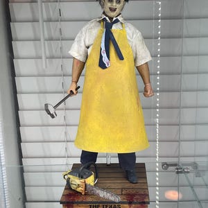 May include: A figure of a character from "The Texas Chainsaw Massacre" movie, wearing a mask, yellow apron, and holding a hammer. A chainsaw and the movie title are on a wooden base. The figure is displayed in a glass case.