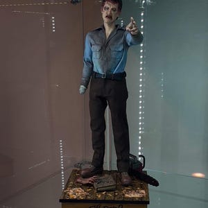 May include: A detailed action figure of a character from Evil Dead 2, wearing a blue shirt and brown pants. The figure is posed with a chainsaw and a book, standing on a wooden base with the film's title. The figure is in a display case.