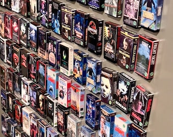 Peut inclure: Un mur recouvert de cassettes VHS de films d'horreur. Les cassettes sont disposées en rangées et en colonnes, et elles présentent des titres tels que "Jeu d'enfant", "Hellraiser", "Les Dents de la mer", "The Thing", "Halloween", "Scream", "Vendredi 13", et "Nightmare on Elm Street".