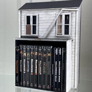 May include: A white model house with a black roof and a black door. The house is on a shelf with a collection of Halloween movie DVDs. The DVDs are all in black cases with the title "Halloween" and a number. The DVDs are arranged in order from 1 to 13.