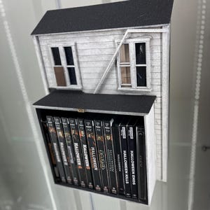 May include: A white and black model of a house with a black roof. The house has two windows and a broken window. The house is sitting on a shelf with a collection of Halloween movie DVDs.