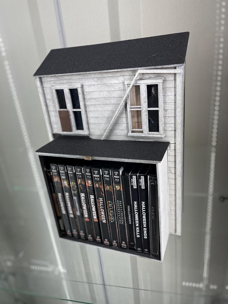 May include: A white and black model of a house with a black roof. The house has two windows and a broken window. The house is sitting on a shelf with a collection of Halloween movie DVDs.