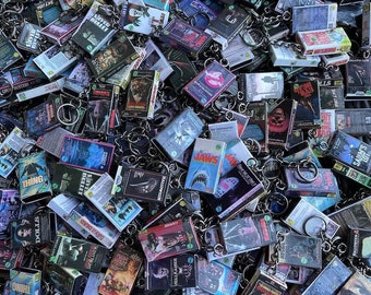 Peut inclure: Un assortiment de porte-clés miniatures en forme de cassettes VHS, représentant des titres de films d'horreur classiques tels que "Jaws", "Halloween" et "The Thing". Les porte-clés sont de couleurs et de designs variés, avec des anneaux argentés.