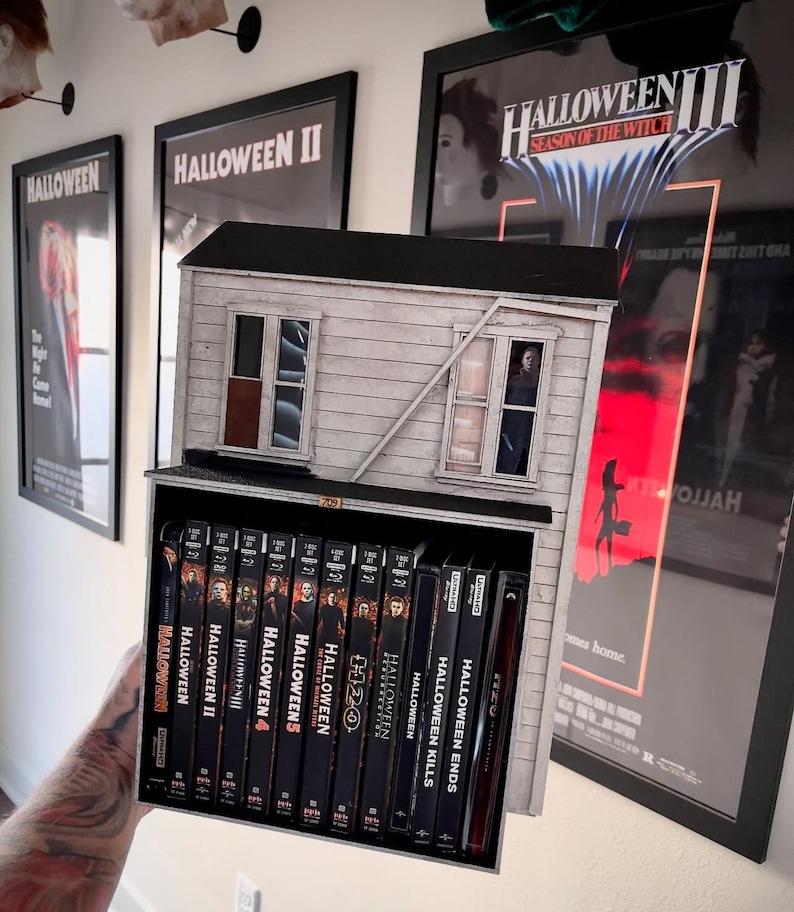 May include: A miniature white house with black trim and windows, holding a collection of Halloween movie DVDs. The DVDs are arranged in a shelf inside the house. The house is in front of a wall with three movie posters for Halloween, Halloween II, and Halloween III: Season of the Witch.