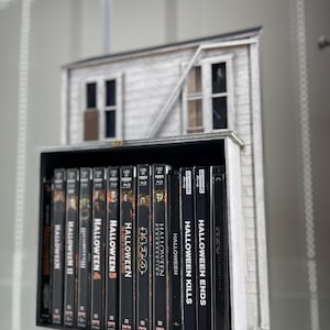 May include: A collection of Halloween movie DVDs, including Halloween Ends, Halloween Kills, Halloween Resurrection, Halloween H20, Halloween 4, Halloween 5, Halloween III, Halloween II, and Halloween. The DVDs are arranged on a shelf in front of a miniature model of a house.