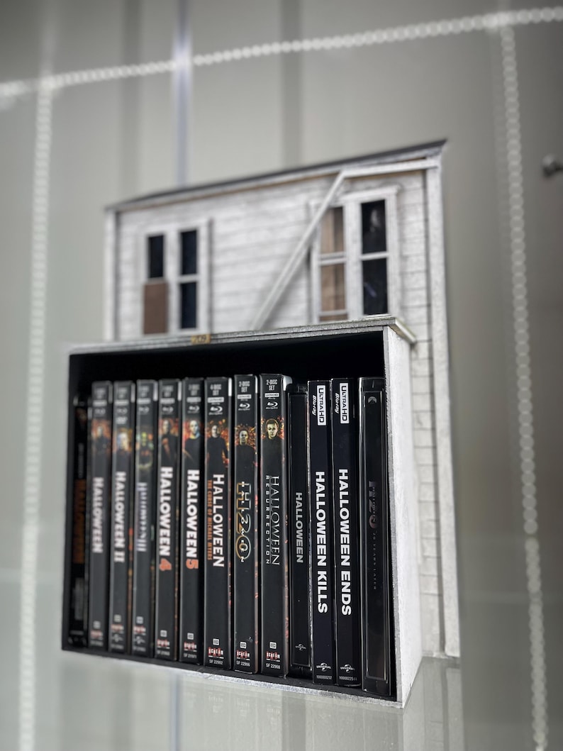 May include: A collection of Halloween movie DVDs, including Halloween Ends, Halloween Kills, Halloween Resurrection, Halloween H20, Halloween 4, Halloween 5, Halloween III, Halloween II, and Halloween. The DVDs are arranged on a shelf in front of a miniature model of a house.