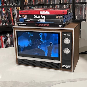 Puede incluir: Un pequeño televisor retro de color marrón que muestra una escena en tonos azules de un personaje con sombrero. Encima hay cuatro cajas de películas: A Nightmare on Elm Street, Child's Play, The Thing y The Lost Boys. El televisor tiene el texto ZOMETH.