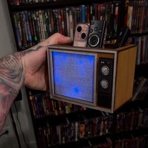 Puede incluir: Un organizador de escritorio de madera con forma de televisor antiguo con una pantalla azul brillante que muestra estática. La parte superior del organizador cuenta con un compartimento abierto que contiene un teléfono inteligente y dos mandos a distancia. El panel frontal incluye dos perillas.