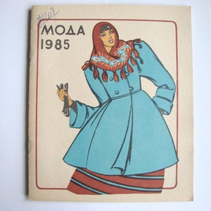 Op de afbeelding: Een vintage tijdschriftcover met een vrouw die een blauwe jas draagt met een rood-witte gestreepte sjaal. De tekst op de cover luidt "MODA 1985".