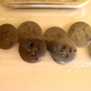 May include: A collection of eight brown buttons with four holes each. The buttons are arranged in a semi-circle on a white surface.