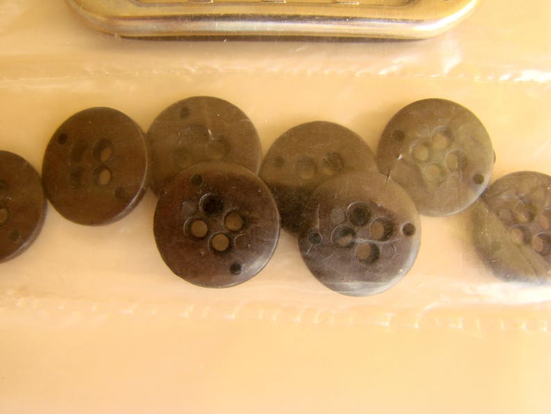 May include: A collection of eight brown buttons with four holes each. The buttons are arranged in a semi-circle on a white surface.