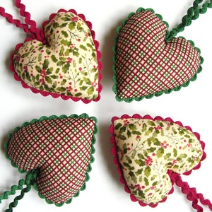 Puede incluir: Cuatro adornos de corazón de tela roja y verde con bordes festoneados. Los corazones están decorados con un patrón de acebo y bayas y un patrón de cuadros.
