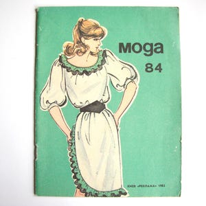 Op de afbeelding: Een vintage naaipatroonboekje met een groene omslag waarop een vrouw is afgebeeld die een witte jurk draagt met een groene en zwarte bies. De titel van het patroon is "Moga 84".