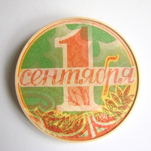 May include: A round, wooden, framed, decorative plate with a green, orange, and red design. The plate features a large, red number "1" and the text "сепmадня" in a stylized font.