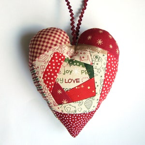 Puede incluir: Un adorno en forma de corazón de patchwork rojo y blanco con las palabras "alegría, paz, amor" impresas en él.