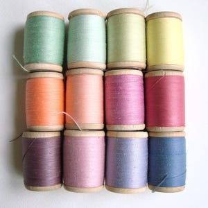 Op de afbeelding: Assortiment van 10 spoelen garen in verschillende pastelkleuren, waaronder roze, blauw, groen, geel en oranje. De spoelen zijn gerangschikt in een rasterpatroon.