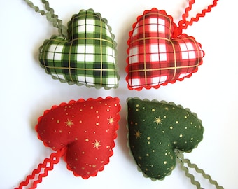May include: Four fabric heart ornaments with different patterns. Two hearts are green and white plaid, one is red and white plaid, and one is red with gold stars. Each heart has a scalloped edge and is attached to a ribbon.