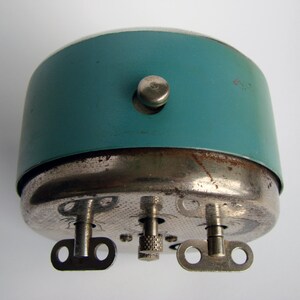May include: A teal blue alarm clock with a silver metal base and three winding keys. The clock has a small knob on the side.