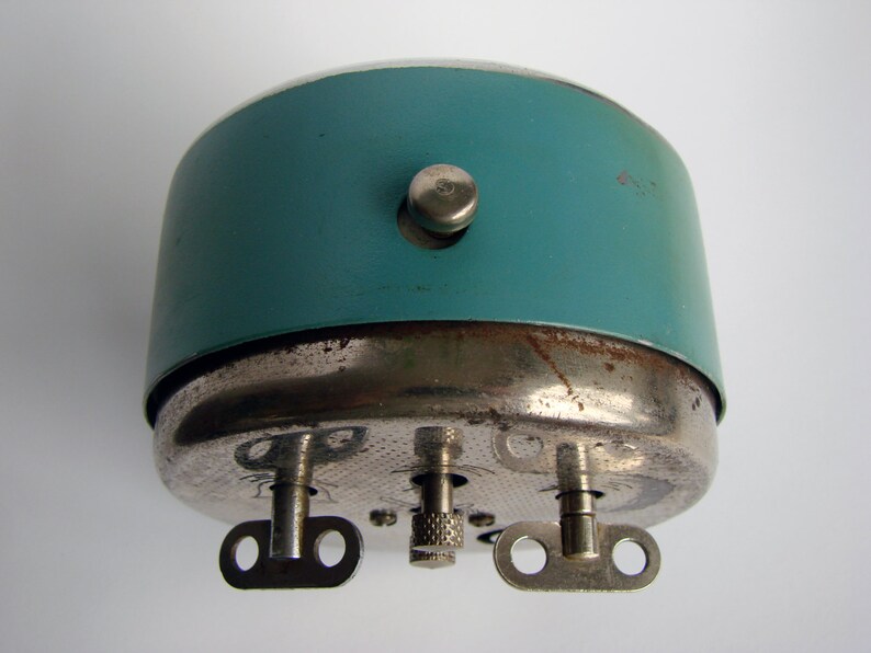 May include: A teal blue alarm clock with a silver metal base and three winding keys. The clock has a small knob on the side.