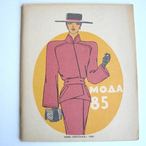 Op de afbeelding: Vintage mode-illustratie uit 1984. De cover toont een vrouw in een roze pak en hoed, met een zwarte clutch en handschoenen. De illustratie staat tegen een gele cirkel met de tekst "MOДA 85".