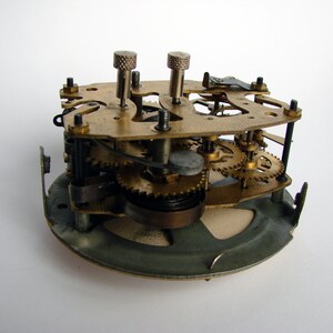 May include: Close-up of a clock mechanism with brass gears and a metal base. The clockwork is exposed, showing the intricate workings of the timepiece.
