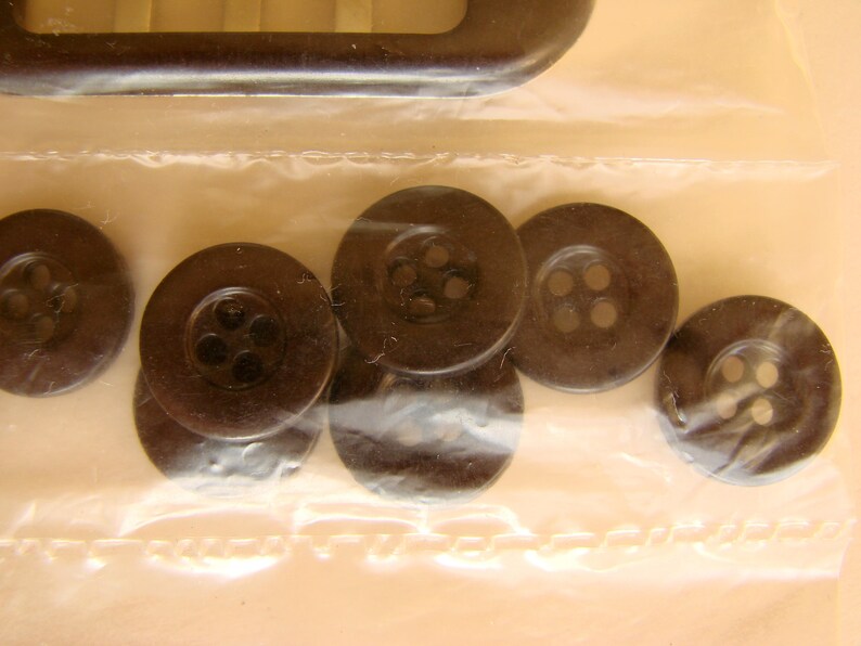 May include: A close-up of six black plastic buttons with four holes each. The buttons are arranged in a semi-circle on a clear plastic surface.