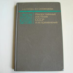 Op de afbeelding: Een grijs hardback boek met de titel "Лекарственные растения СССР и их применение" in gouden letters. Het boek heeft een groene en witte illustratie op de voorkant.