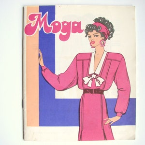 Op de afbeelding: Een vintage tijdschriftcover met een vrouw in een roze jurk met een witte kraag en strik. De titel van het tijdschrift is "Moga" in een gestileerd lettertype.