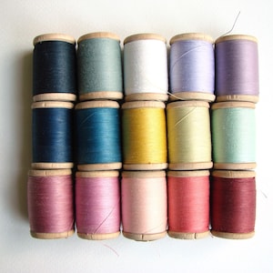 Op de afbeelding: Een verzameling van vijftien houten klossen naaigaren gerangschikt in een raster van drie rijen en vijf kolommen. De kleuren van het garen zijn zwart, saliegroen, wit, lavendel, paars, marineblauw, blauwgroen, mosterdgeel, lichtgeel, munt, roze, lichtroze, koraal en donkerrood.