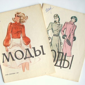 Op de afbeelding: Twee vintage modebladen met het woord "Моды" gedrukt op de omslagen. Het blad links toont een vrouw in een rood blouse en een witte rok. Het blad rechts toont twee vrouwen in jassen.