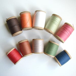 Può includere: Assortimento di dieci bobine di filo di vari colori, tra cui rosso, arancione, marrone, verde, blu e bianco. Le bobine sono in legno e hanno un aspetto vintage.