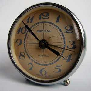 May include: A vintage silver alarm clock with a cream-colored face. The clock has black hands and blue numbers. The clock face also has the text "SEVANI" and "4 JEWELS" printed on it. The clock is made in the USSR.