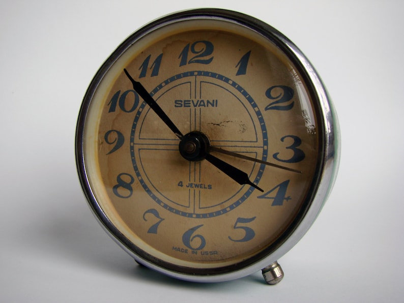 May include: A vintage silver alarm clock with a cream-colored face. The clock has black hands and blue numbers. The clock face also has the text "SEVANI" and "4 JEWELS" printed on it. The clock is made in the USSR.